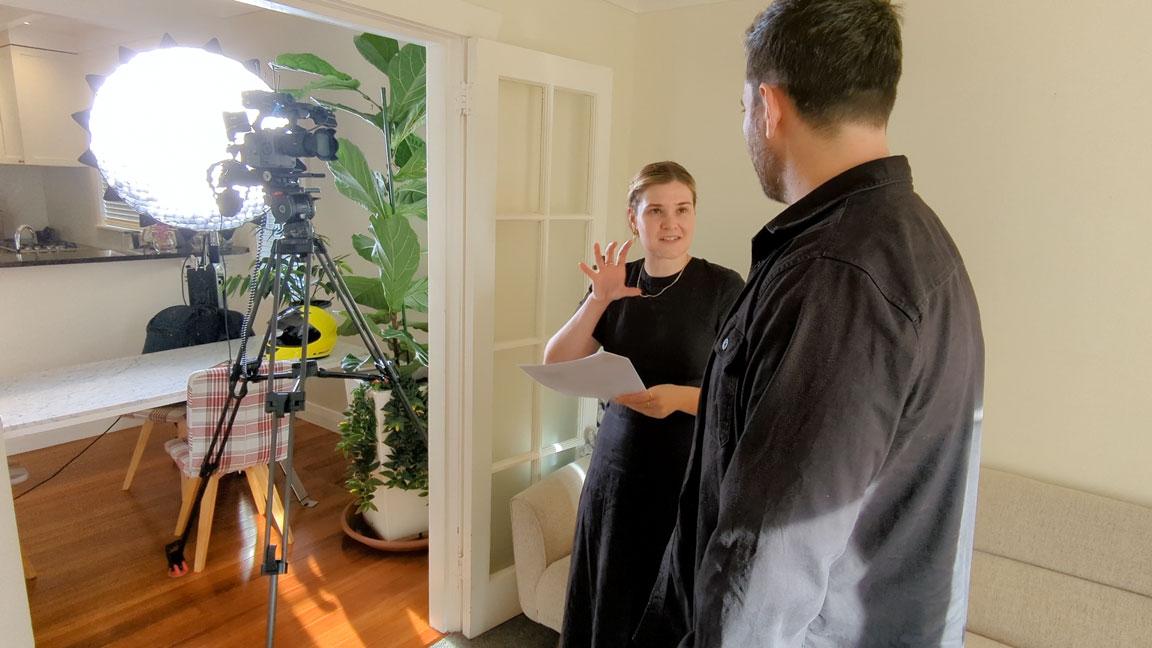 A man and woman in the process of video production standing in front of a camera in a living room.